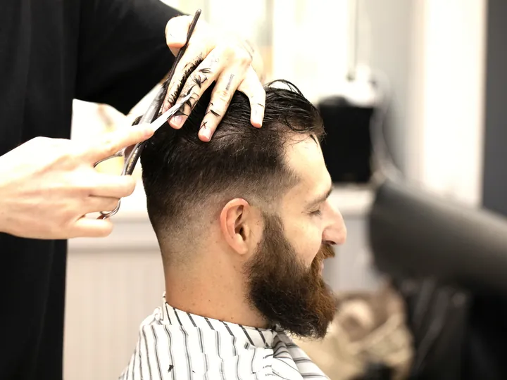 A stylist cutting a seated man's hair with scissors and comb.