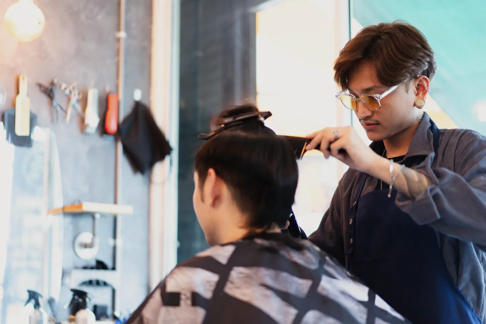 A stylist trimming a seated client's hair in a small neighbourhood salon, tools hanging on the wall behind.