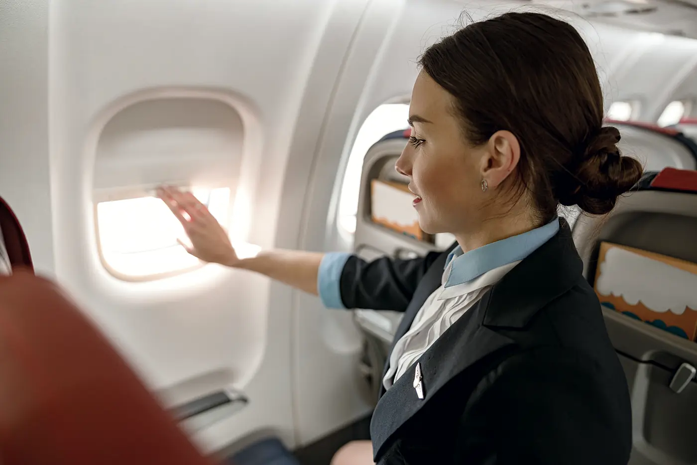 A cabin host, in profile, lowering a window shade with one hand.