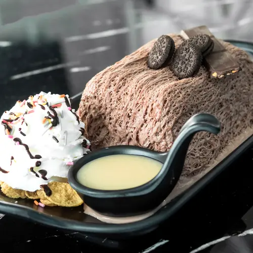 Chocolate bingsu on a dark plate with a jug of condensed milk beside it.