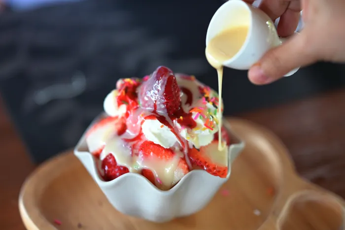 Condensed milk being poured from a small jug over a bowl of shaved ice and strawberries.