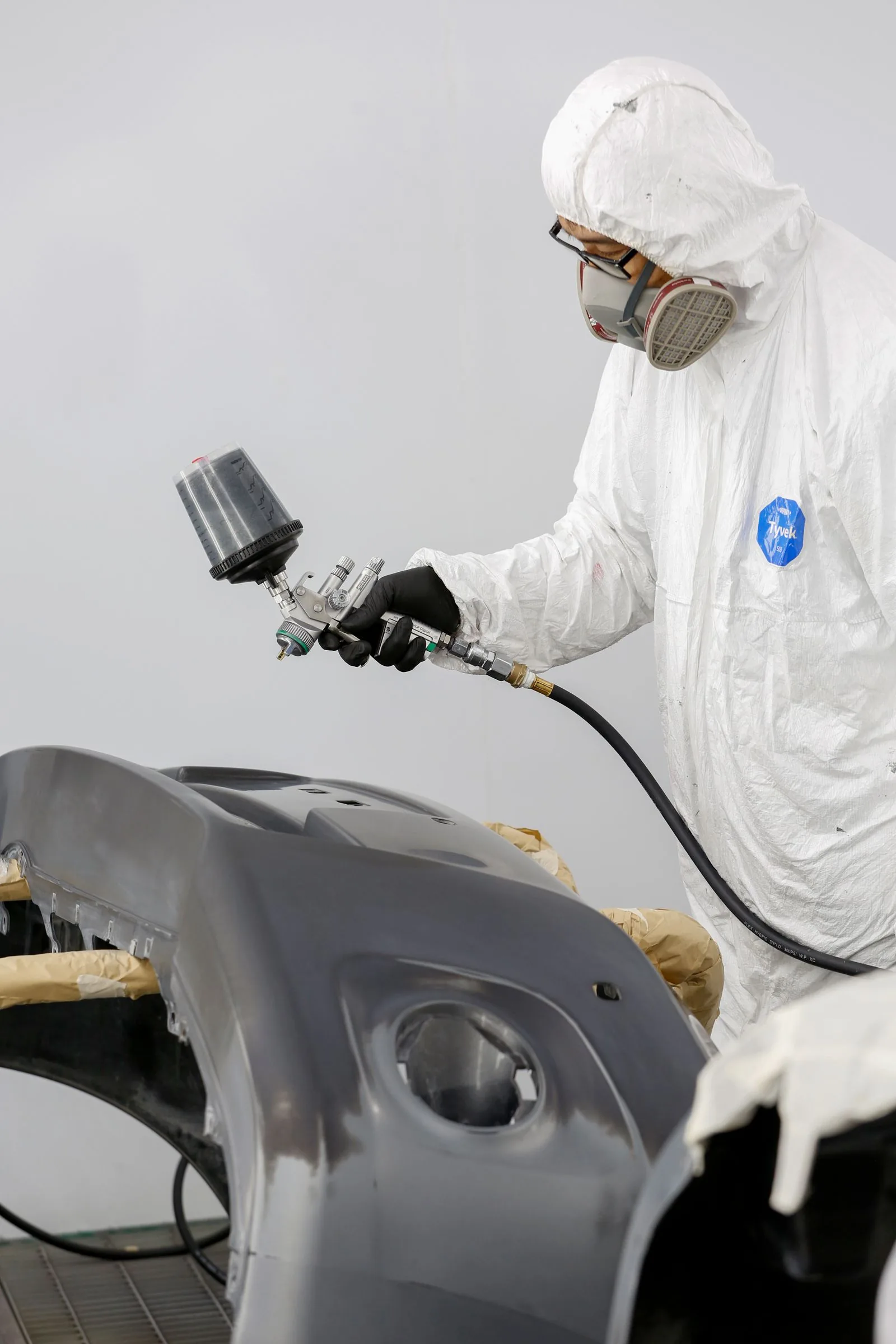 Technician in a protective suit spray-applying coating to a car bumper in a clean booth