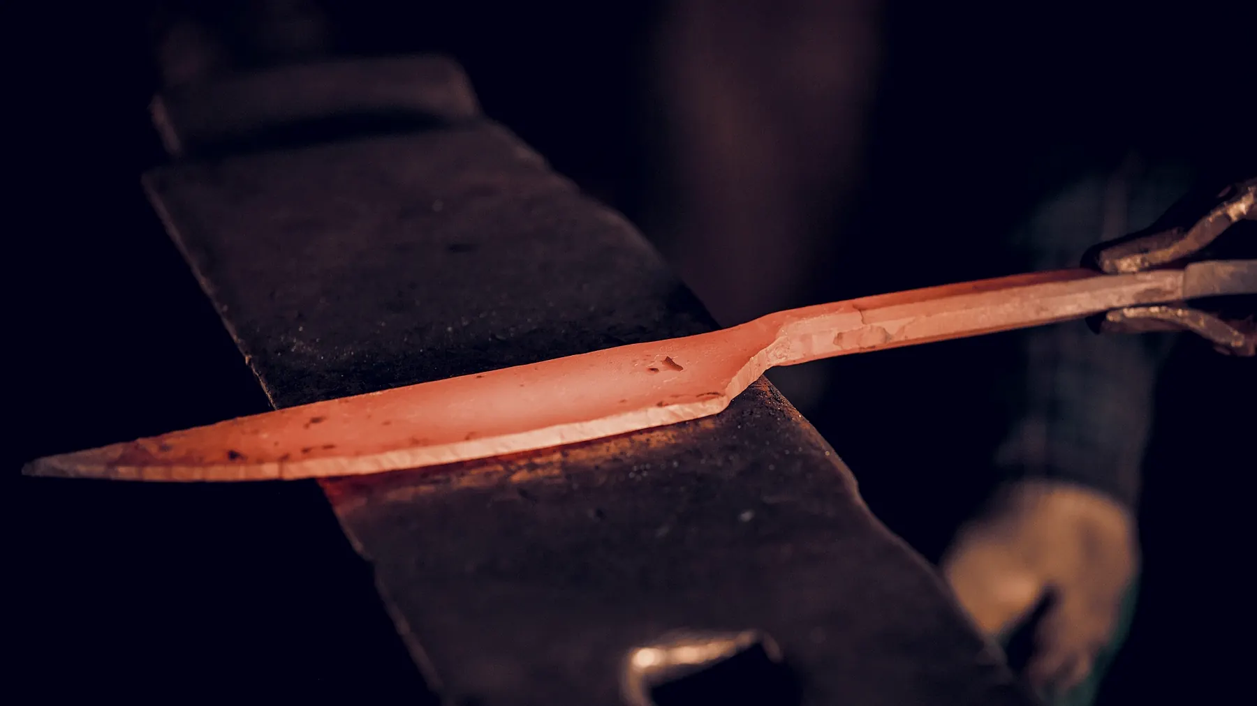 A knife blank at orange heat lying across the face of an anvil, held in a pair of tongs.