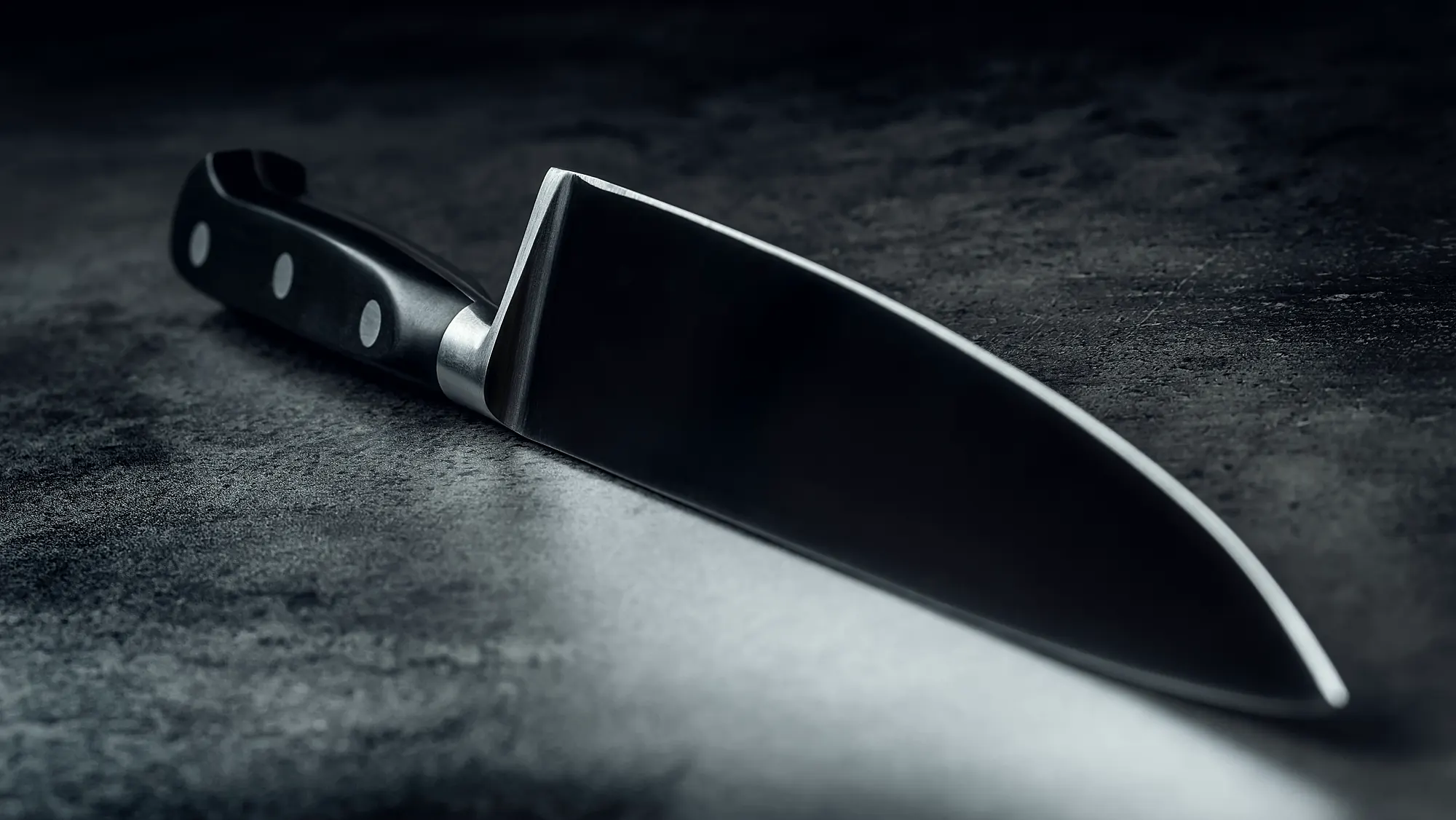 A finished kitchen knife lying on a dark stone bench, the edge catching a single low light.
