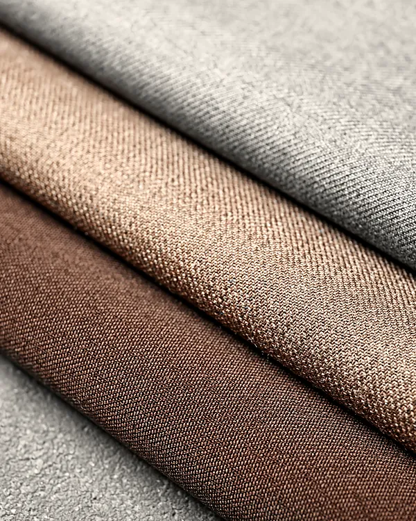 Three bolts of woollen cloth rolled on a bench: a grey twill, a camel and a walnut brown.