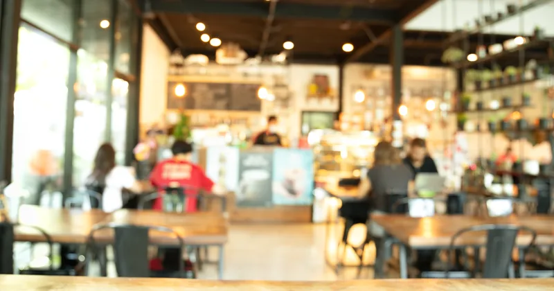 A busy cafe interior seen across the room, the counter and customers softly out of focus.