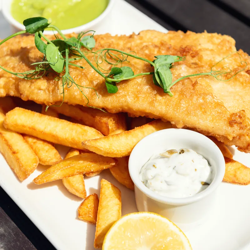 Battered fish with thick-cut chips, tartare sauce and a lemon wedge on a white plate.