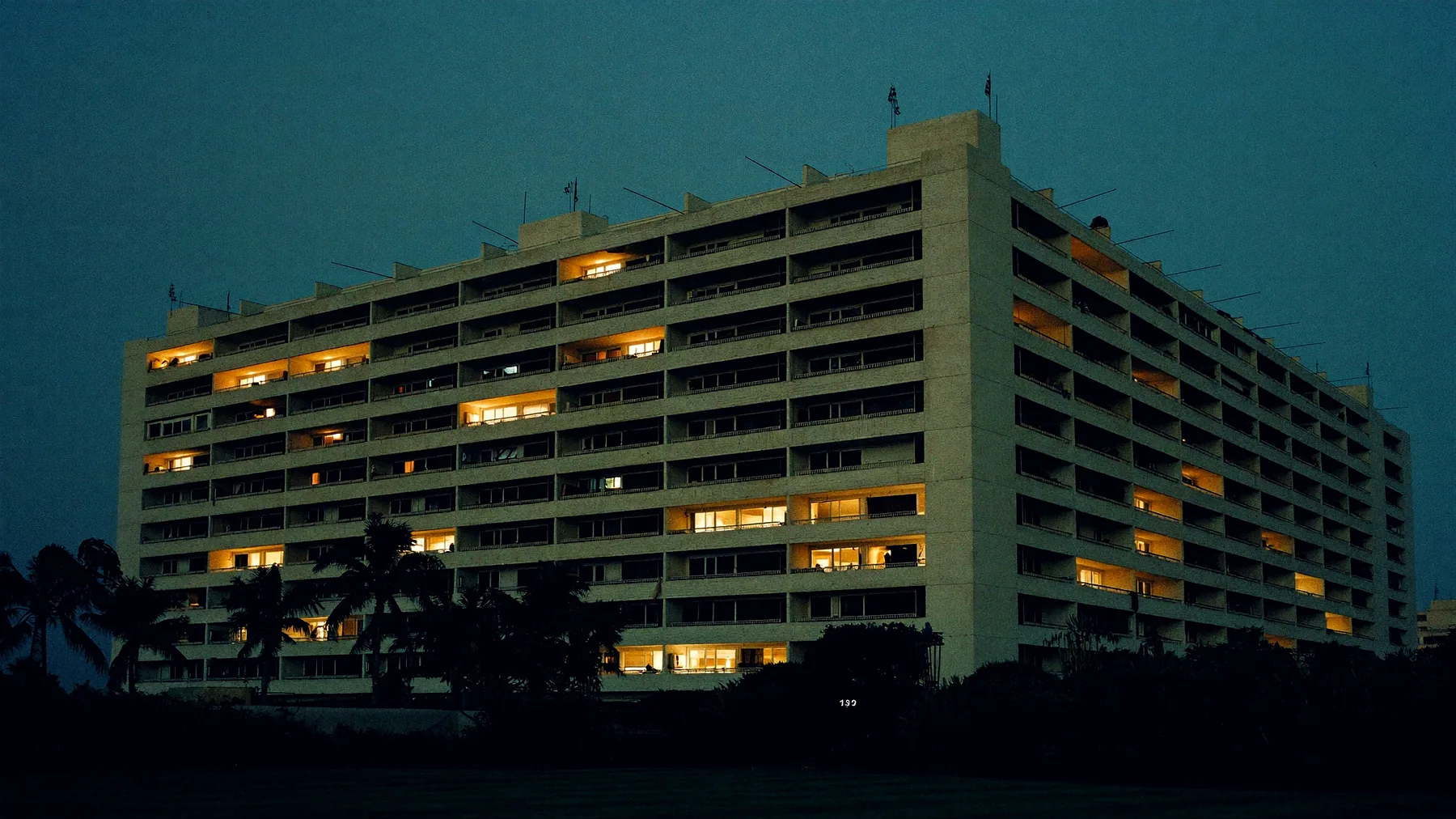 A long public housing block at dusk seen across the estate grass, deep recessed balconies and hundreds of windows, about half of them lit warm amber.