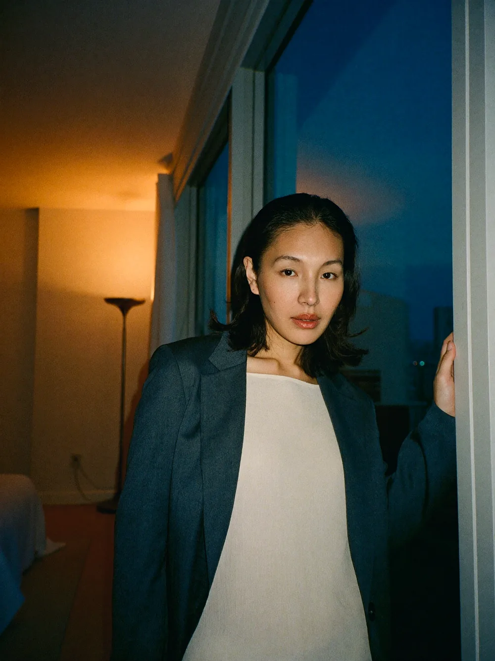 Mabel Seow standing beside the window of an empty living room at dusk, one hand on the window frame, lit by a lamp out of frame.