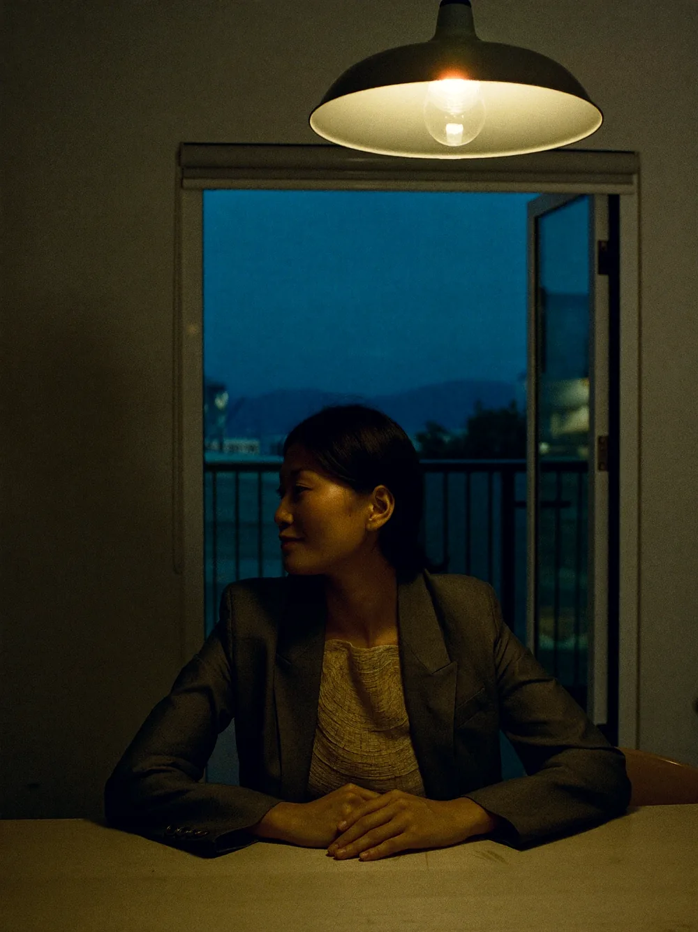 Mabel Seow seated at the end of a bare wooden dining table in an empty flat, looking toward the window under a single pendant lamp.