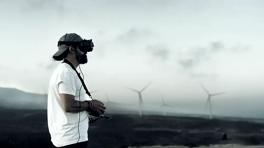 A pilot in goggles, in profile, transmitter in both hands, wind turbines behind.