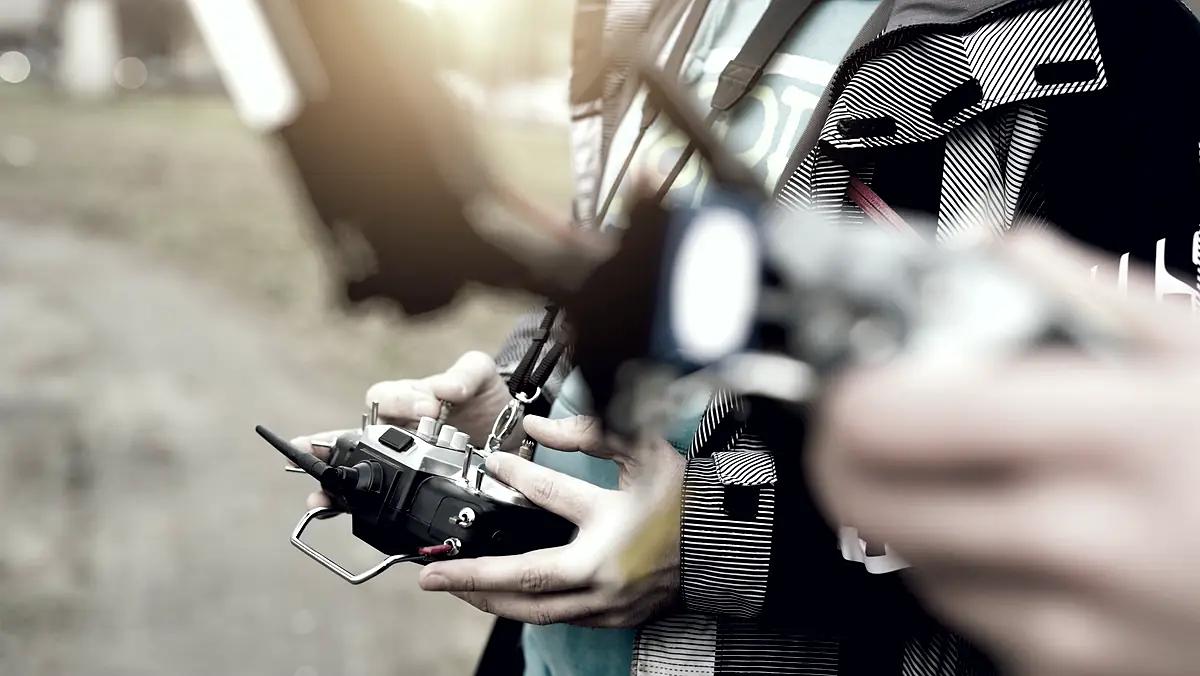 A pilot's hands on a transmitter, thumbs on the sticks, a second pilot blurred beside them.
