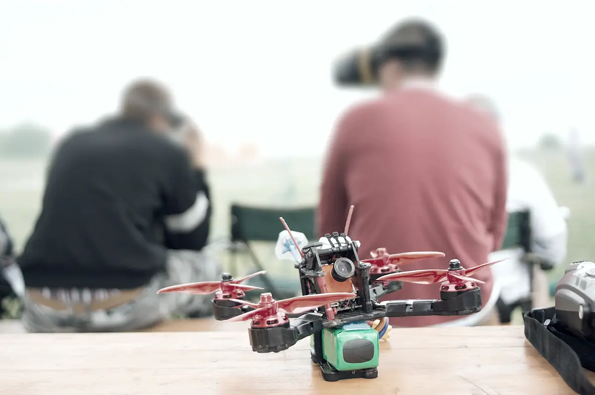 A red racing frame on a trestle table, and behind it three pilots in goggles with their backs to the camera.