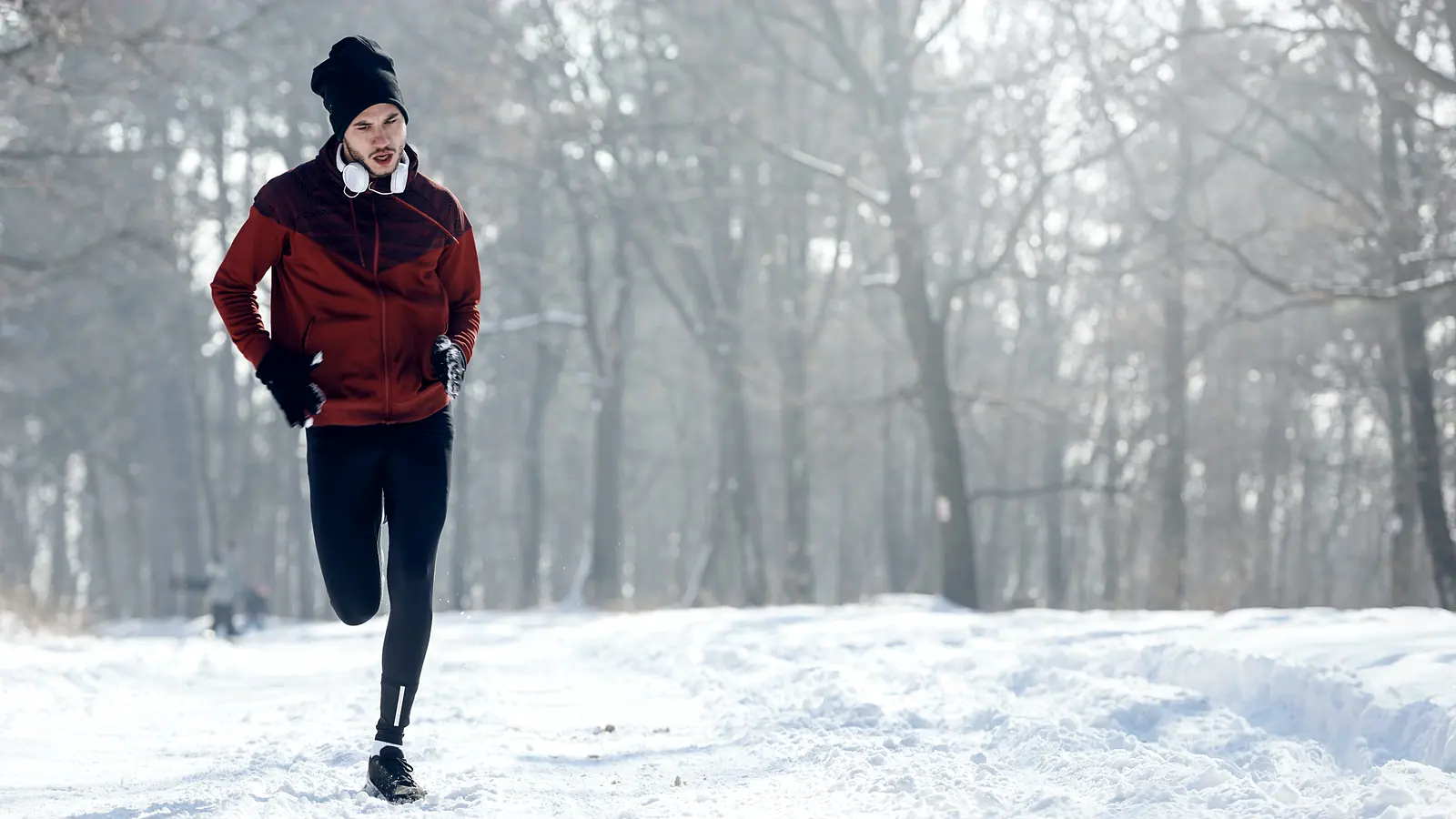 A runner in a hooded winter jacket and tights coming towards the camera along a snow-covered path through bare trees.