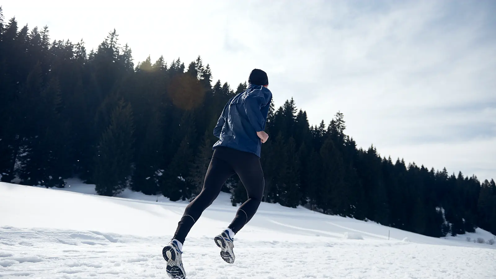 A runner in a shell jacket and tights crossing deep snow below a pine forest.