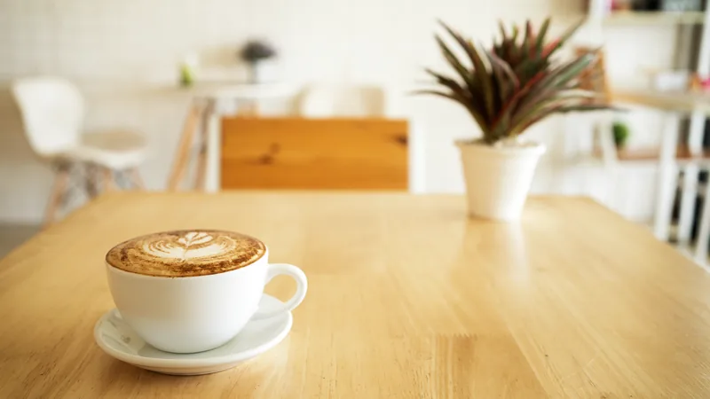 A cappuccino in a white cup on a light wooden table in a bright café.