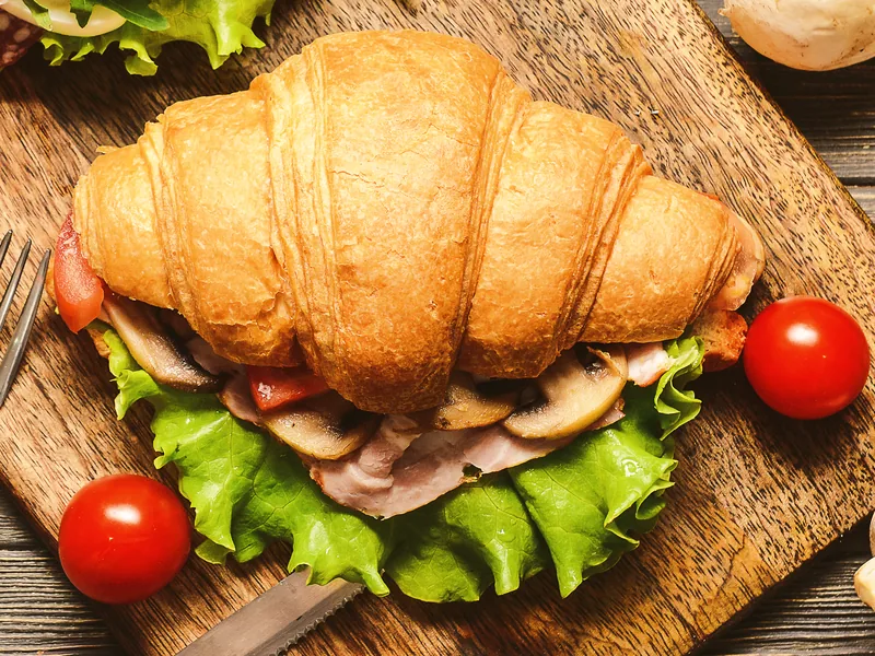 A croissant split and filled with ham, mushrooms and lettuce on a wooden board.
