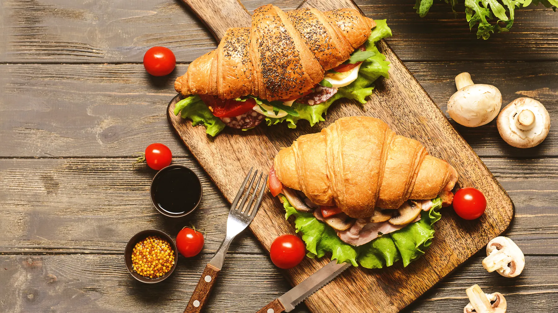 Two filled croissants on a wooden board with cherry tomatoes, mushrooms and lettuce, photographed from above.