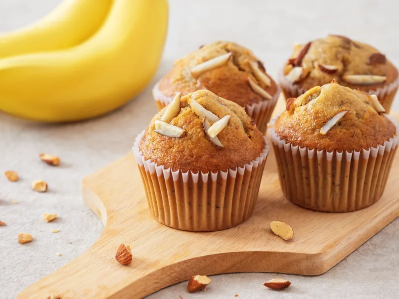 Four banana muffins topped with almond flakes on a wooden board.