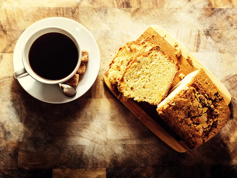 A sliced loaf cake on a wooden board beside a cup of black coffee.