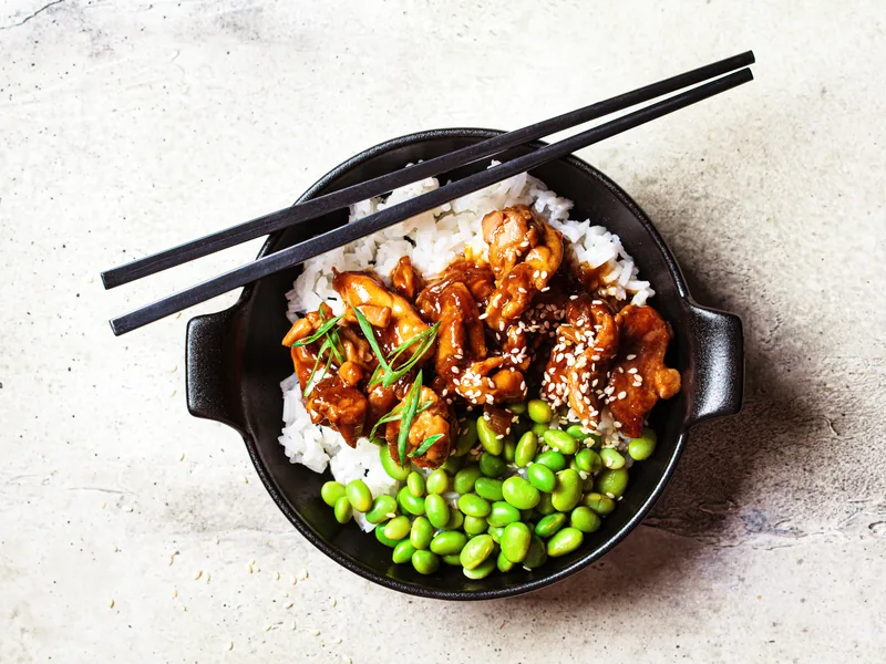 A rice bowl with glazed chicken, green edamame beans and chopsticks resting on the rim.